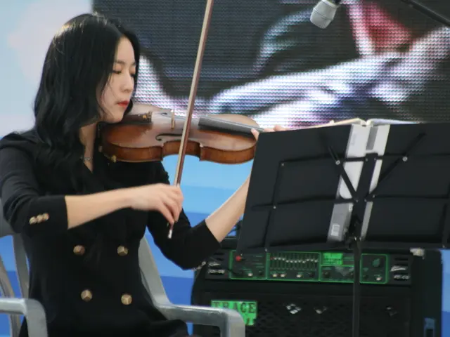 A girl is sitting on a chair wearing a black dress and playing violin. There are papers and its...