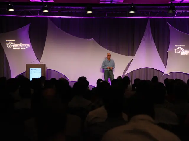 In this picture there is a man wearing blue color shirt, standing on the stage and giving a speech....