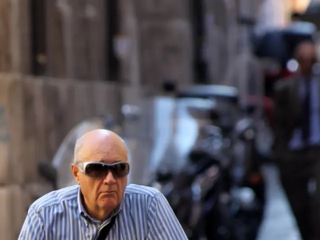 In this image an old man is riding bicycle. He is wearing sunglasses and striped shirt. In the...