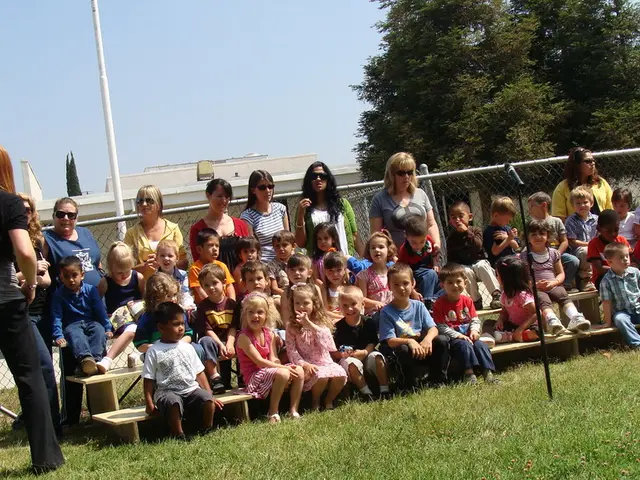 In the center of the image there are children sitting on benches. There are people standing. In the...