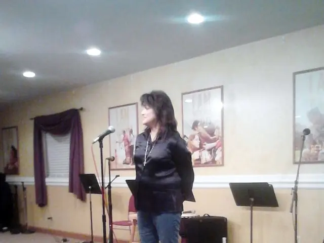 A woman is standing and speaking in the microphone behind her there are photographs on this wall.